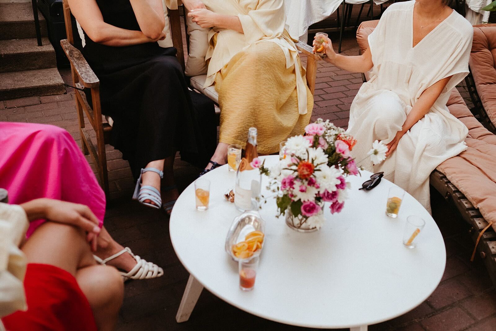 A group of friends wearing flowy dresses gather in chairs around a coffee table outdoors. Small cocktail glasses are set on the coffee table, which has a large floral centerpiece in the middle.