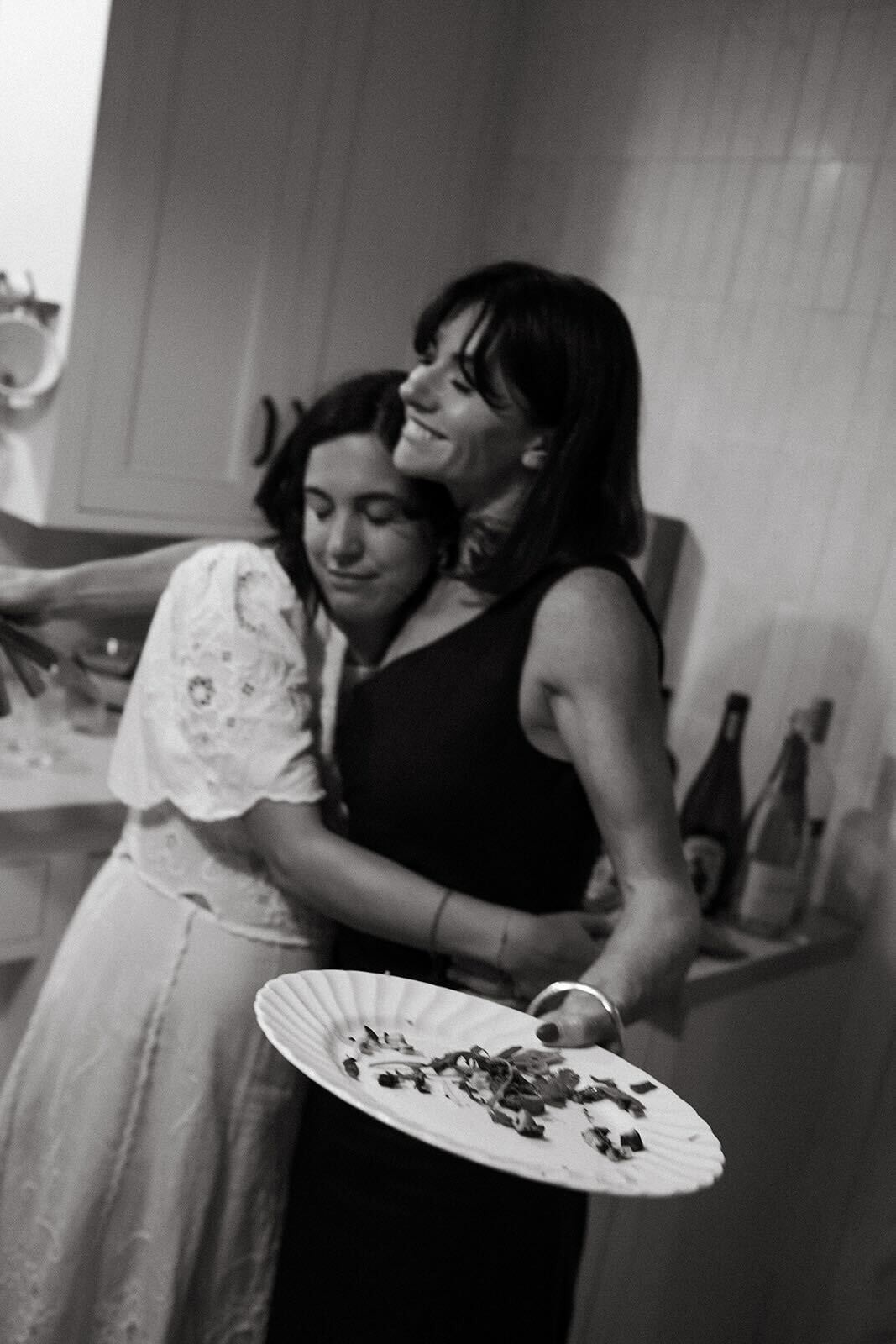 A black and white photo shows two friends hugging and smiling while cleaning up plates after dinner.