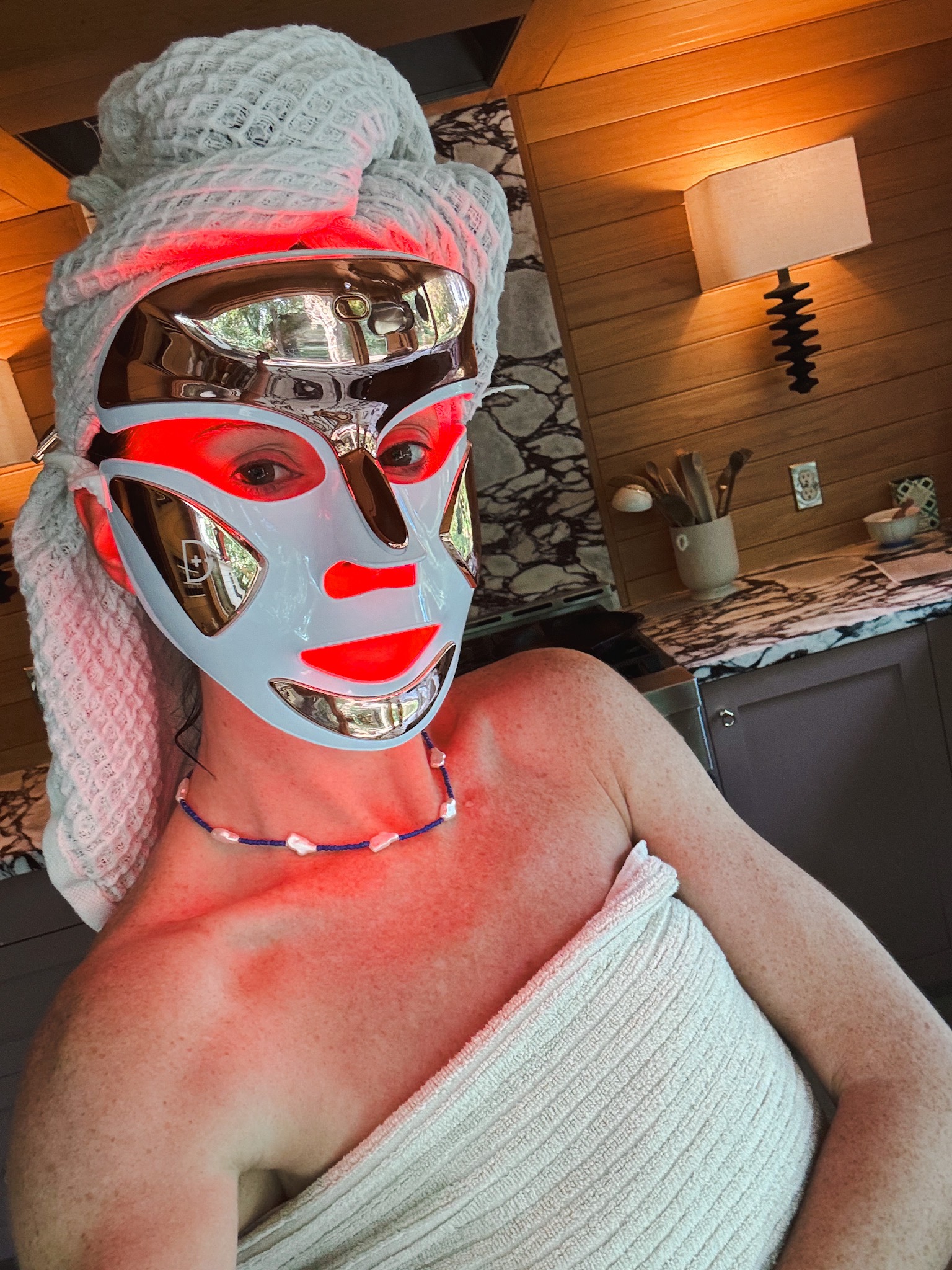 A woman stands in her kitchen wearing the Dr. Dennis Gross LED mask, which is powered on in the red light anti-aging mode.