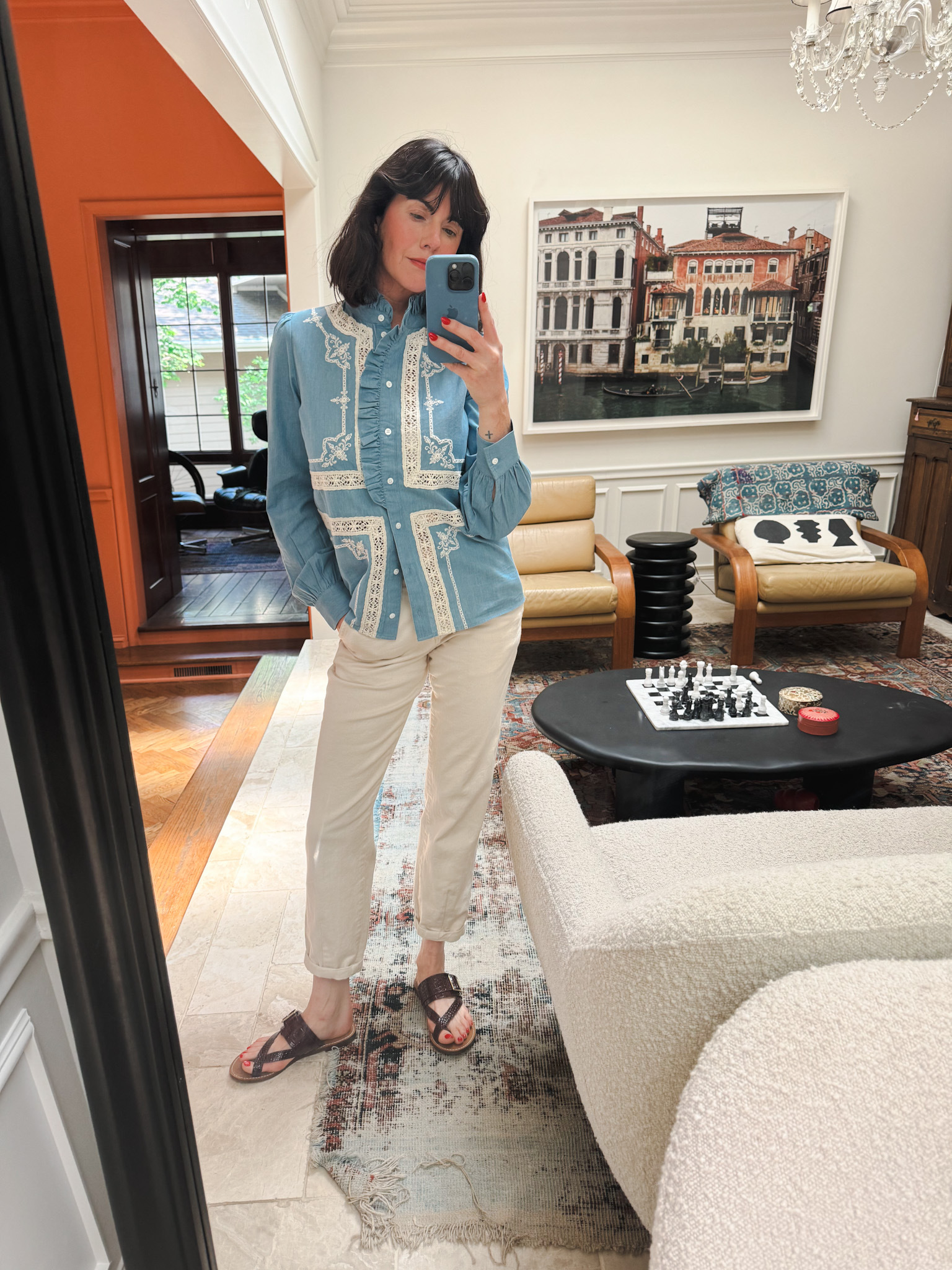 Woman wearing an embroidered chambray blouse, trousers, and sandals taking a mirror selfie.