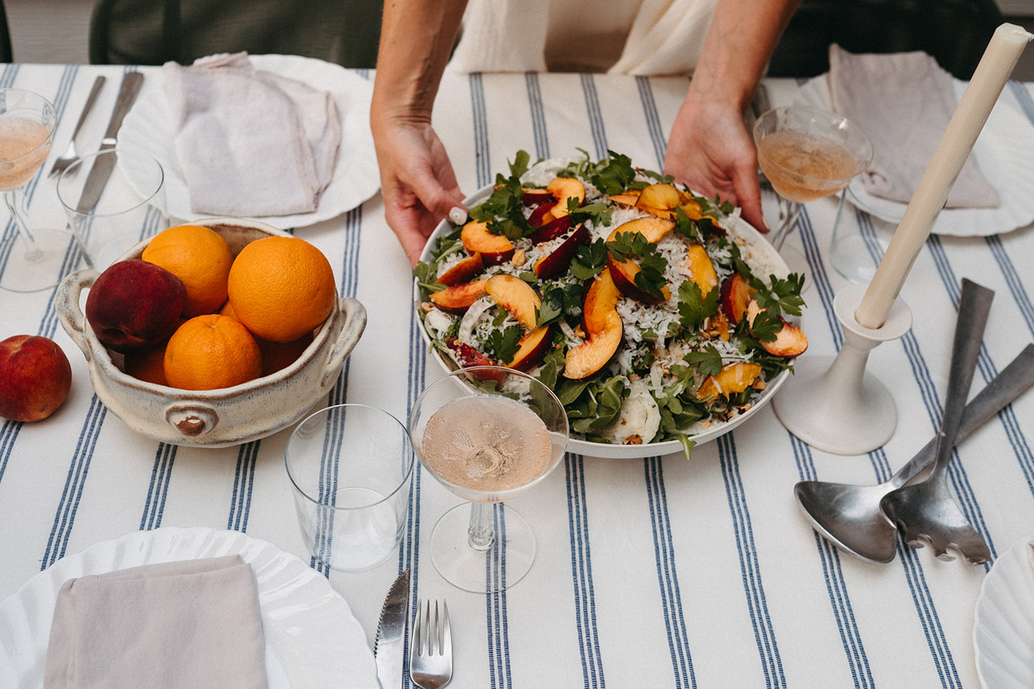 An arugula, fennel, and peach salad is being placed on a tabletop. On the table is a striped tablecloth, tapered candles, peaches and oranges, and glasses of rose.