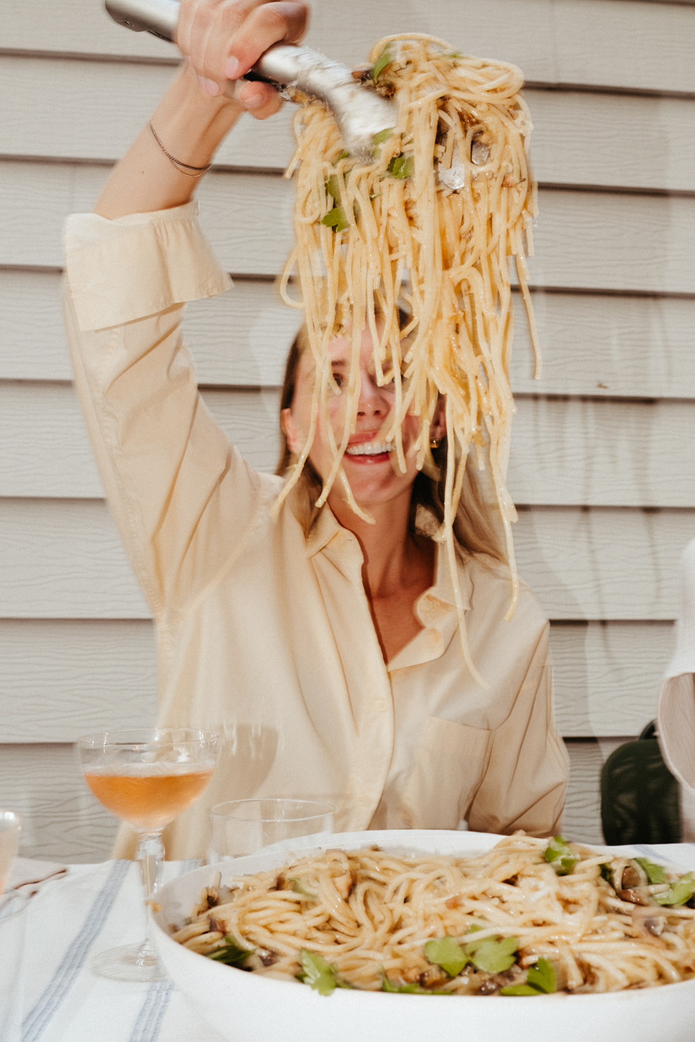 A woman is sitting at an outdoor dinner table using tongs to pick up a large serving of fresh mushroom pasta