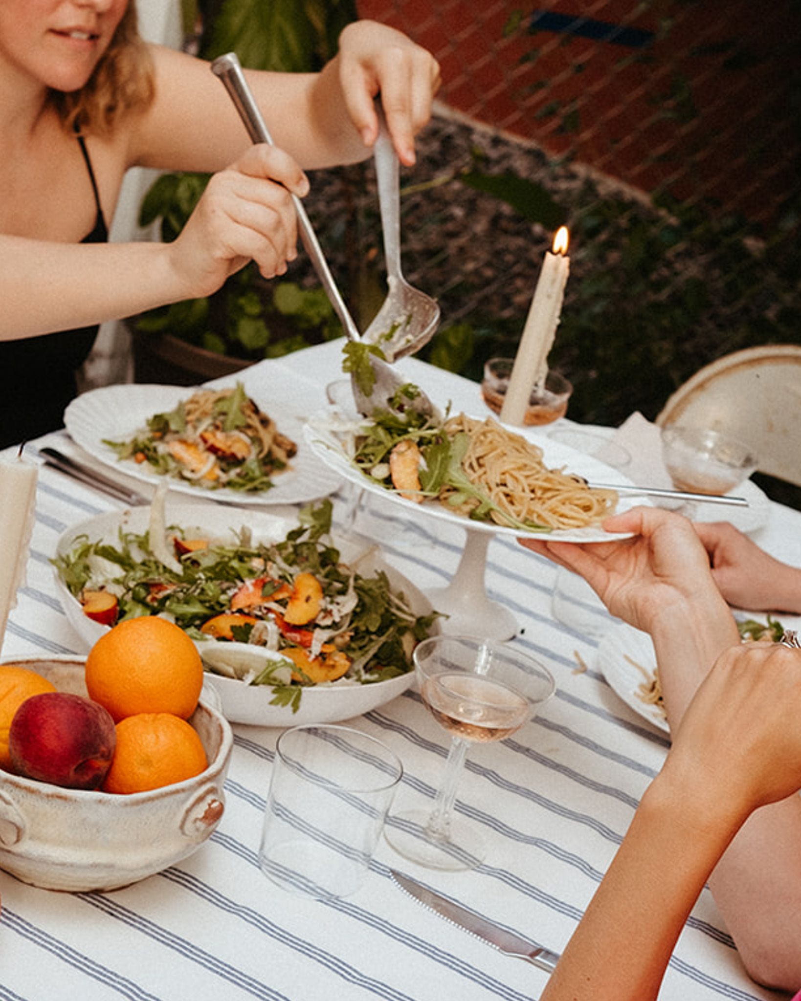 At a casual dinner party outdoors, two women are serving themselves from large bowls of fresh pasta and salad.