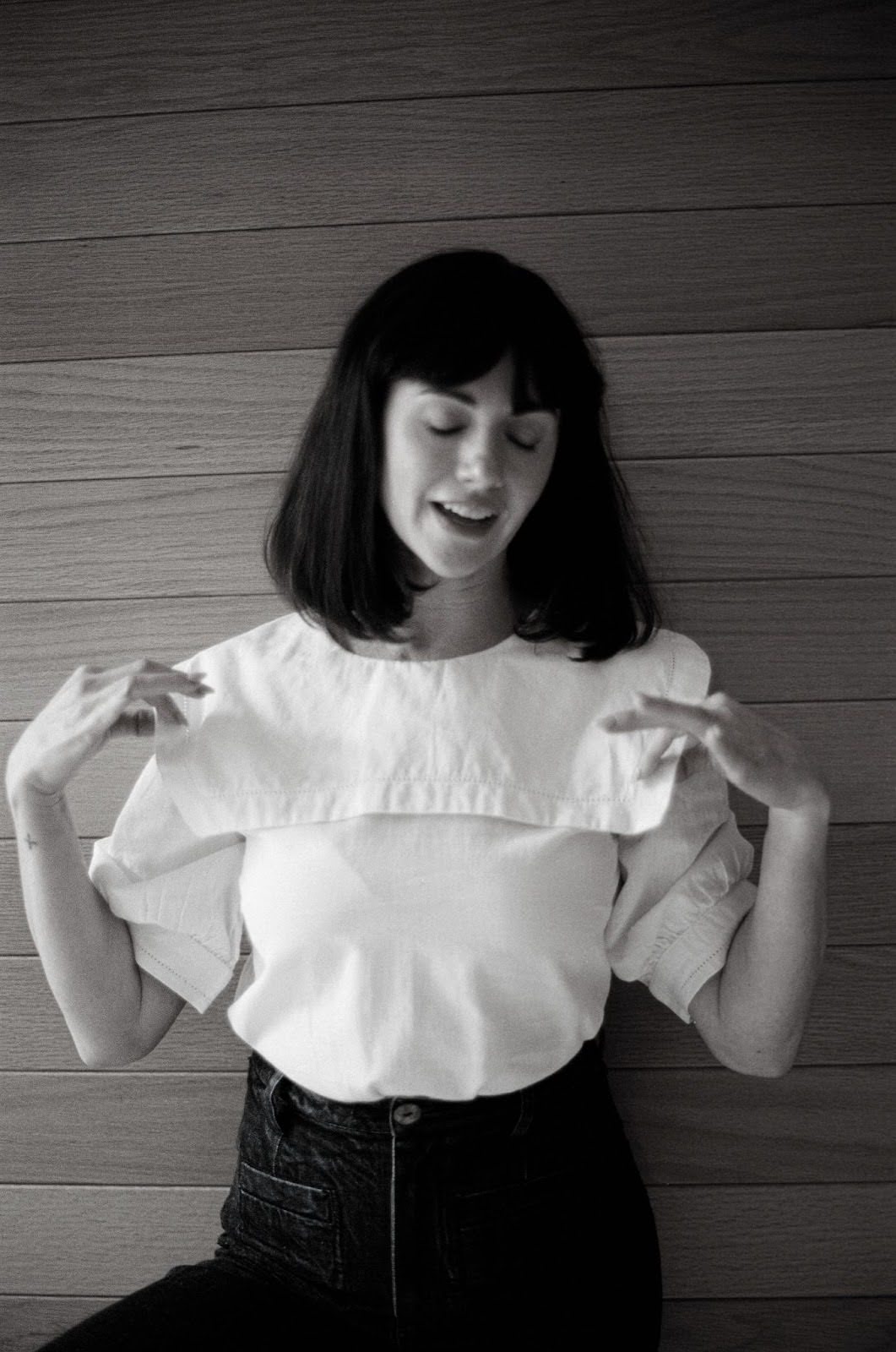 A woman is standing in front of a wood paneled wall, wearing high-waist jeans and a white blouse with puffy sleeves and a sailor collar