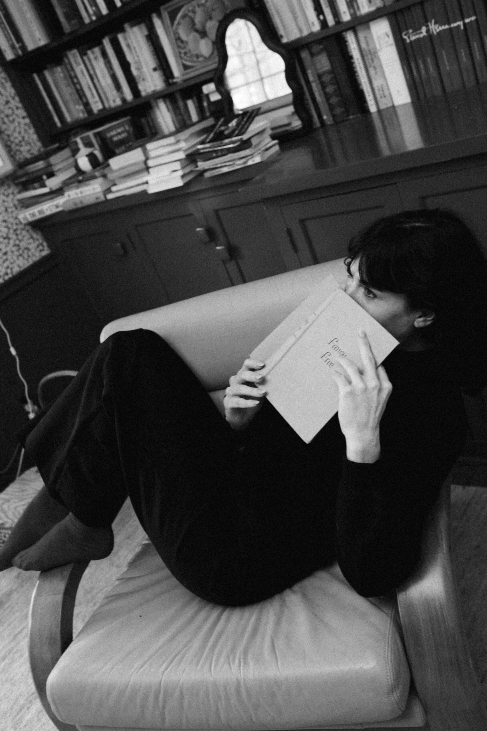 A woman is dressed in all black and sitting in a chair in her office reading a book.