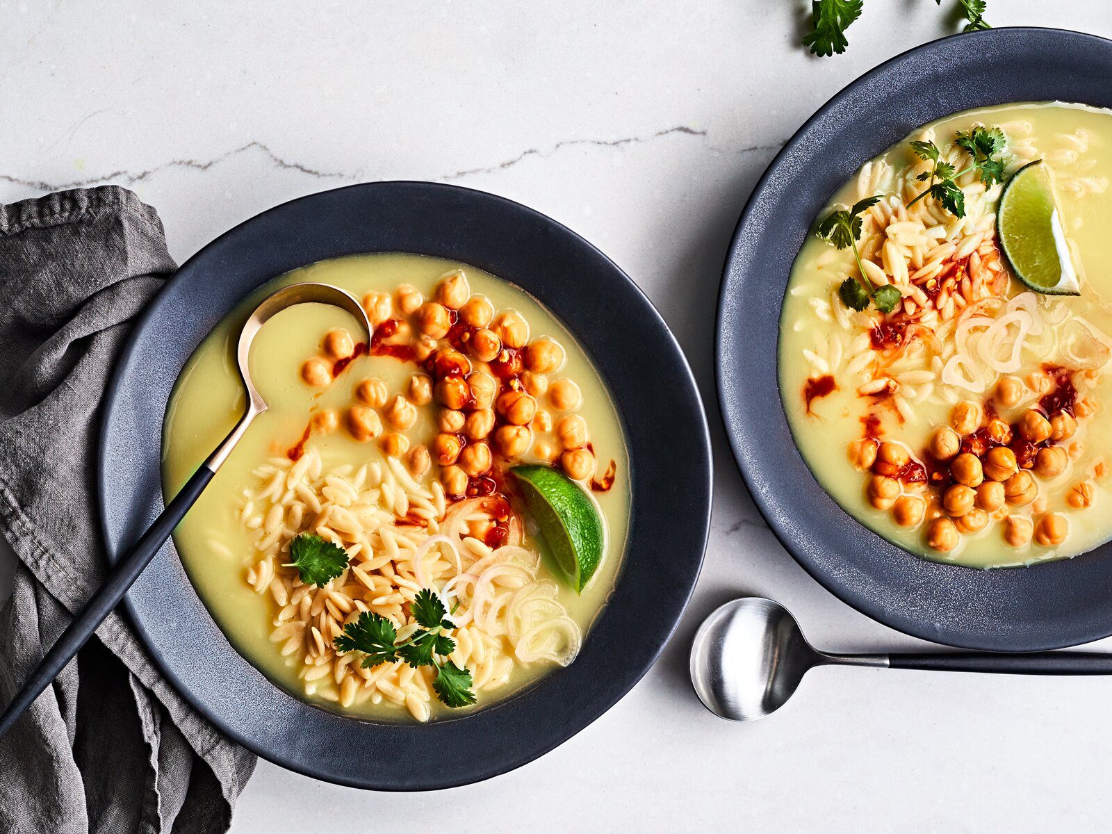 Two bowls of orzo and chickpea soup sit on a marble tabletop