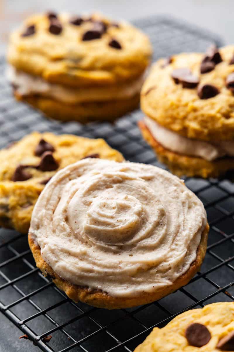 A close up view of a pumpkin chocolate chip cookie topped with a cream filling, next to other pumpkin sandwich cookies.