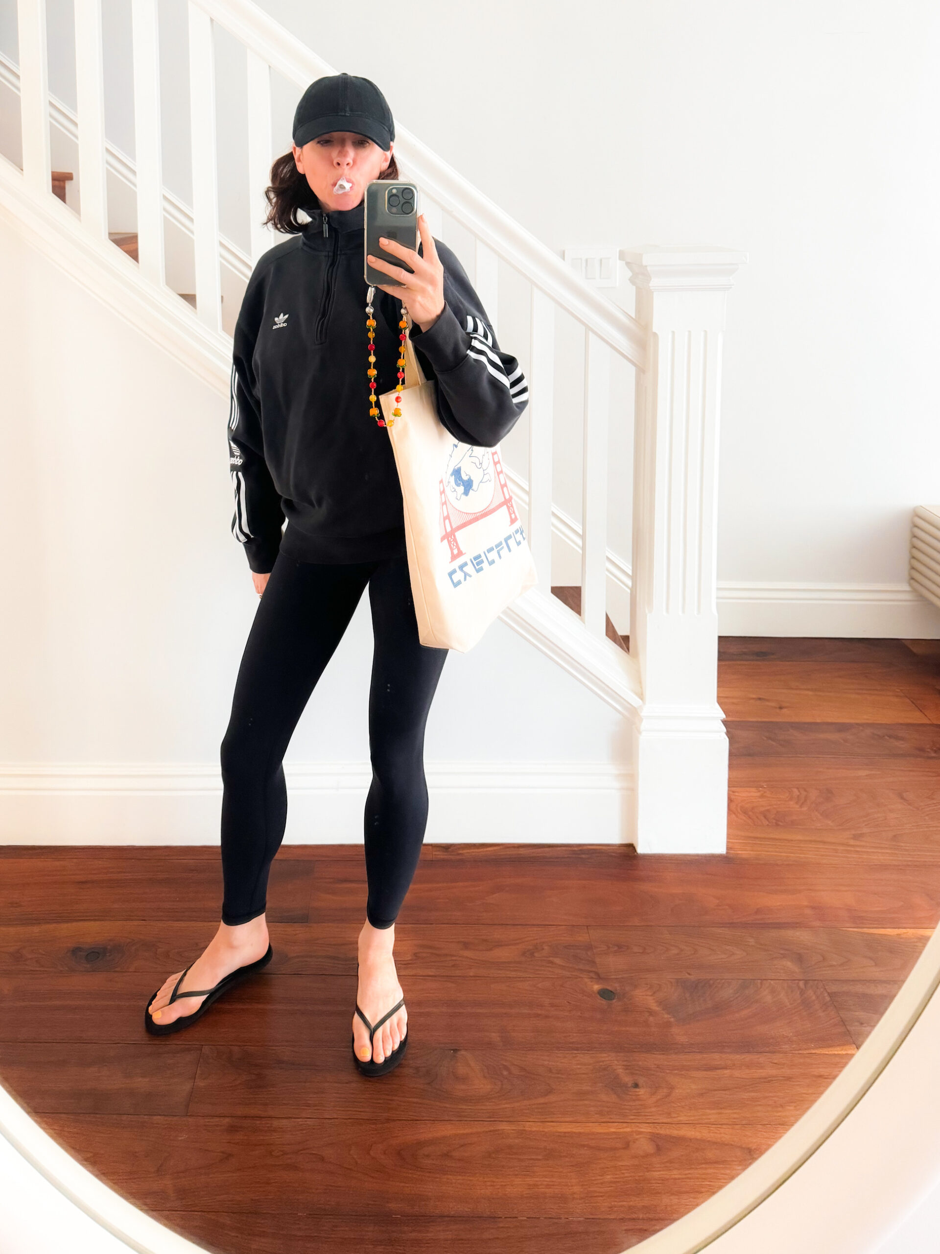 A woman stands in front of a staircase wearing a black half-zip Adidas pullover, black leggings, black flip flops, a black baseball cap, and a canvas tote bag with a graphic of the Golden Gate Bridge on it.