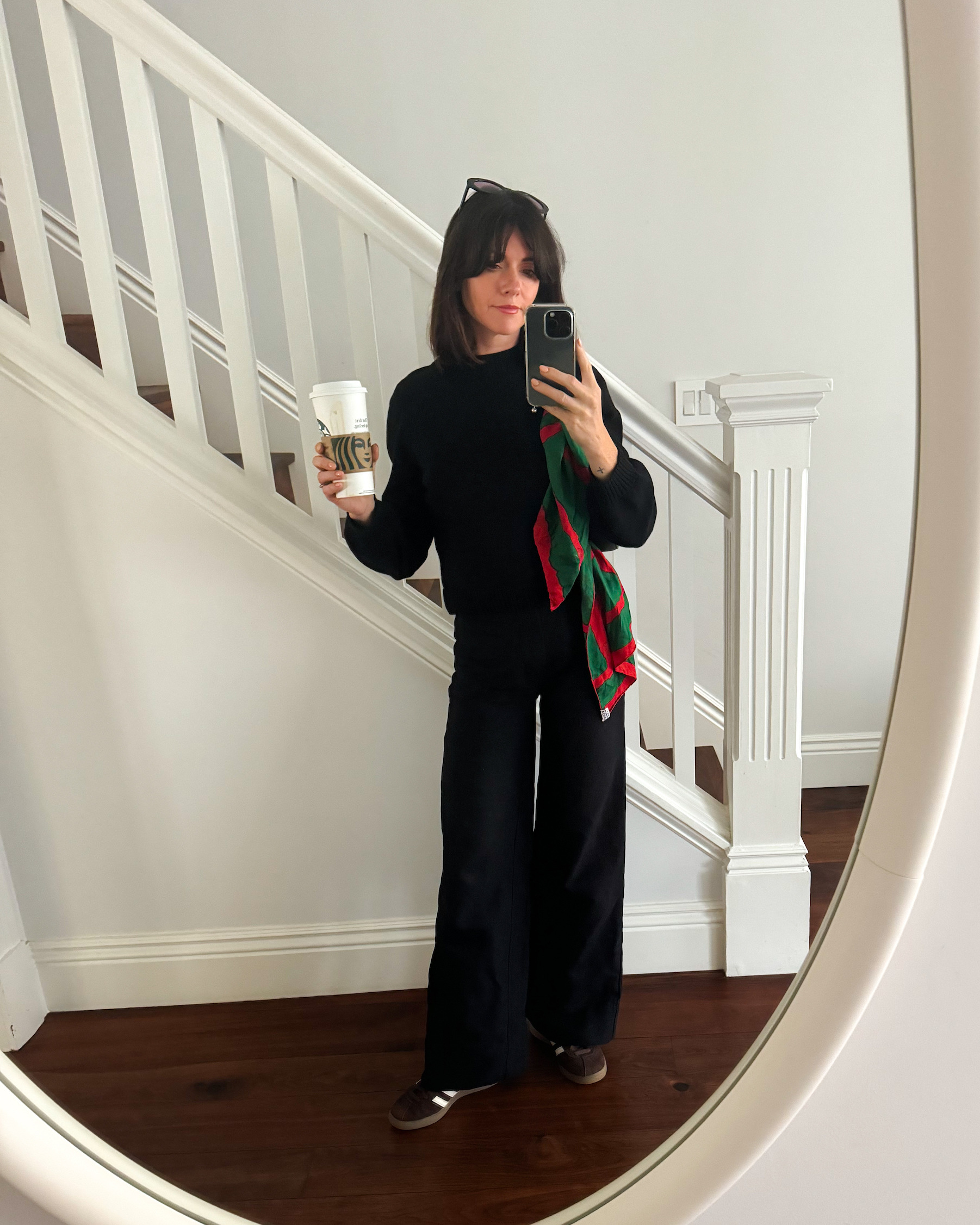 A woman stands in front of a staircase wearing a black sweater, black trousers, brown suede sneakers, and a black shoulder bag with a red and green scarf tied on its handle. She holds a venti Starbucks cup in her hand.