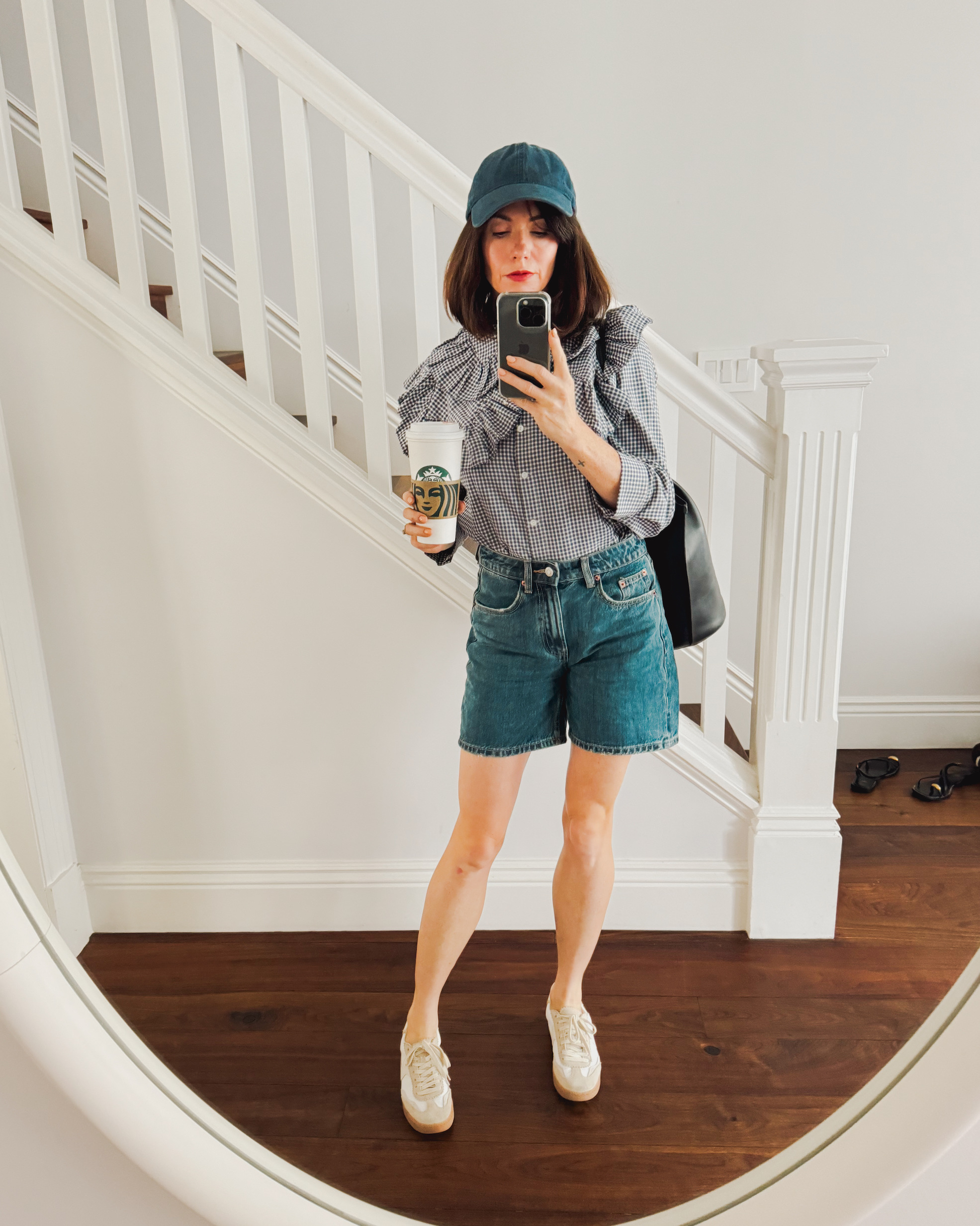 A woman stands in front of a staircase wearing a gingham blouse with an oversized collar, blue denim shorts, ivory sneakers, a blue baseball cap, and a black tote bag. She holds a venti Starbucks cup in her hand.