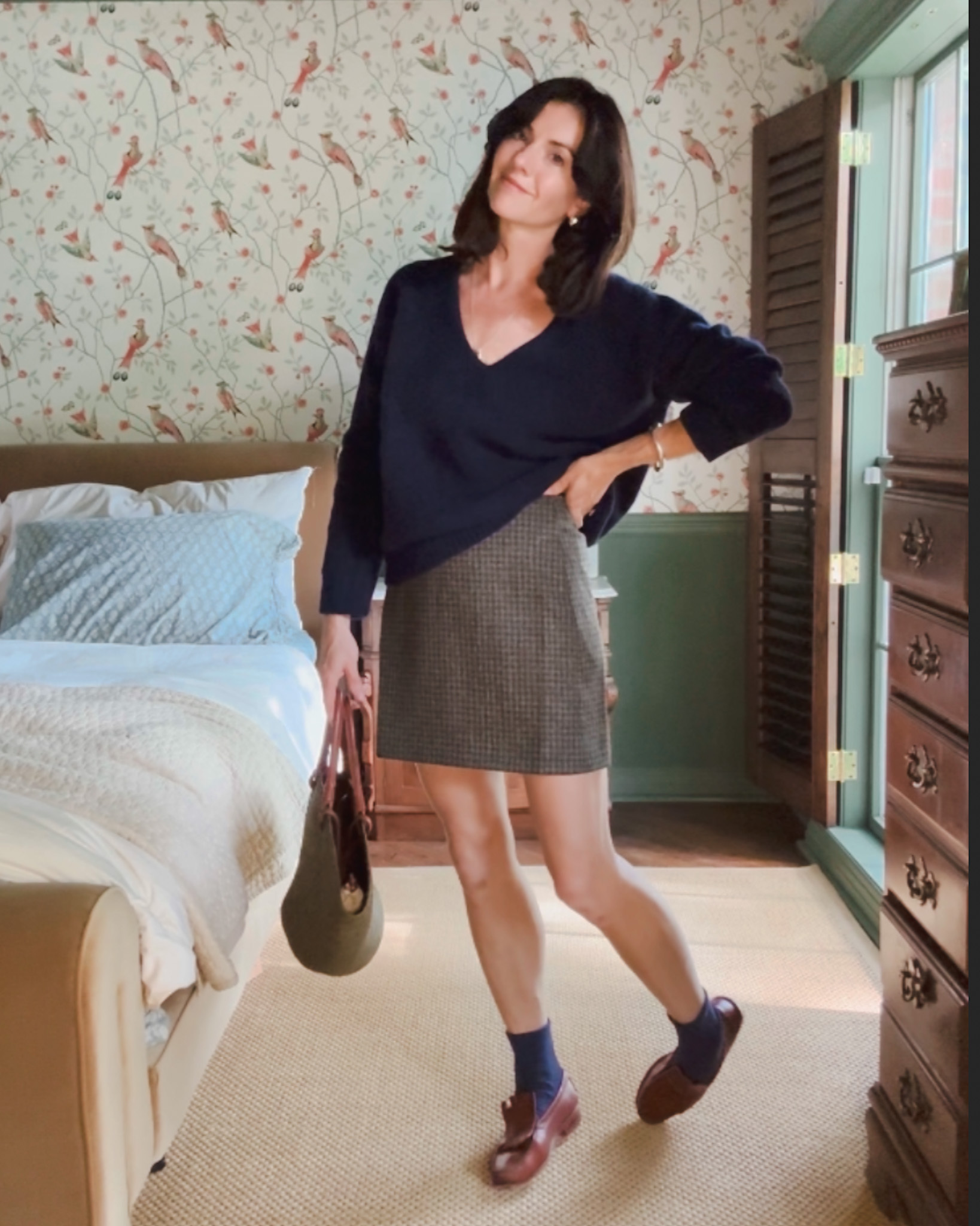 A woman stands in her bedroom wearing pieces from her fall capsule wardrobe: a dark v-neck sweater, a herringbone skirt, navy blue socks, and brown loafers.