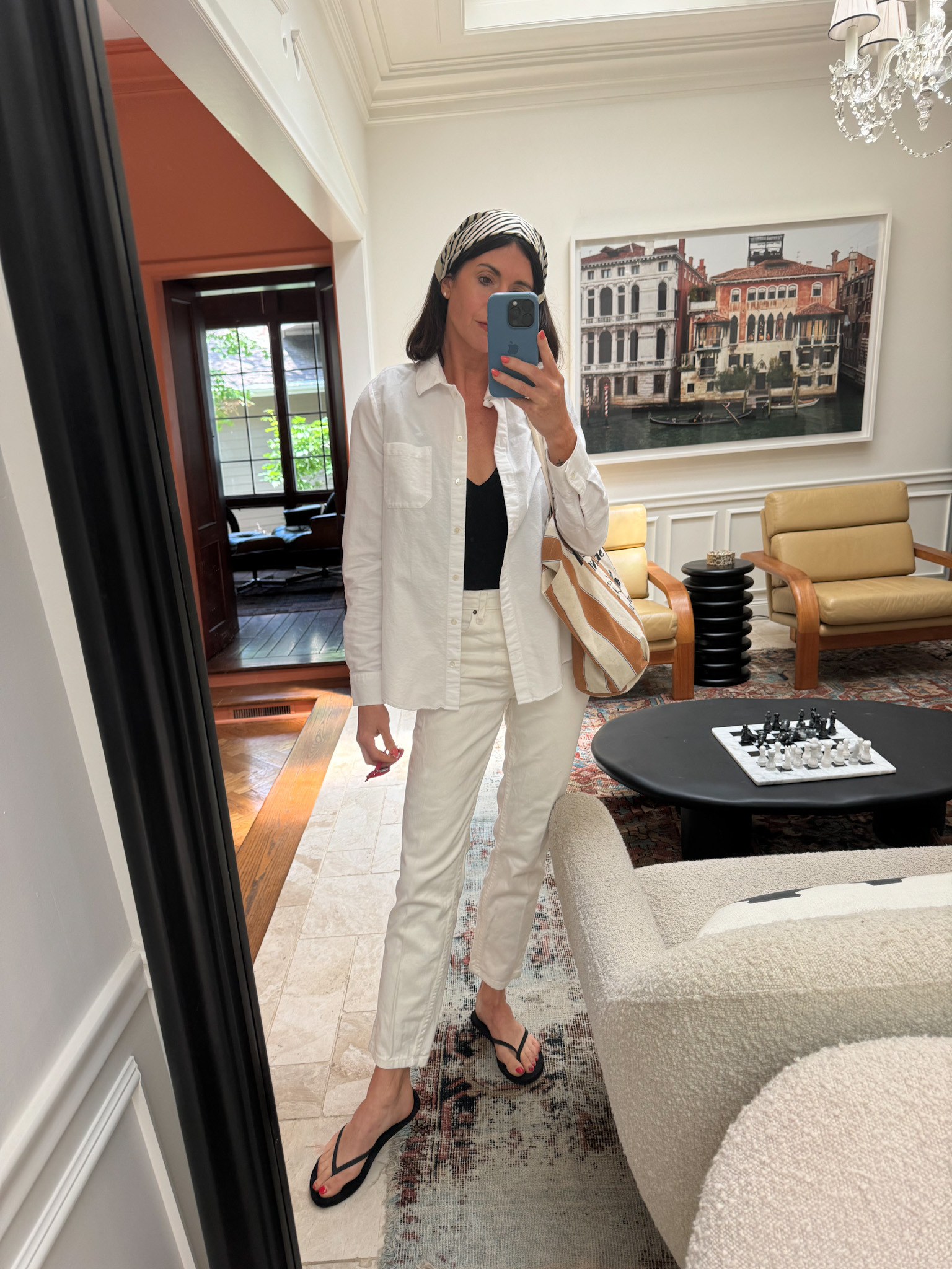 In one of her effortless summer outfits, a woman wears cream-colored jeans, a white button-up, a black swimsuit, black flip flops, and a black and white headscarf