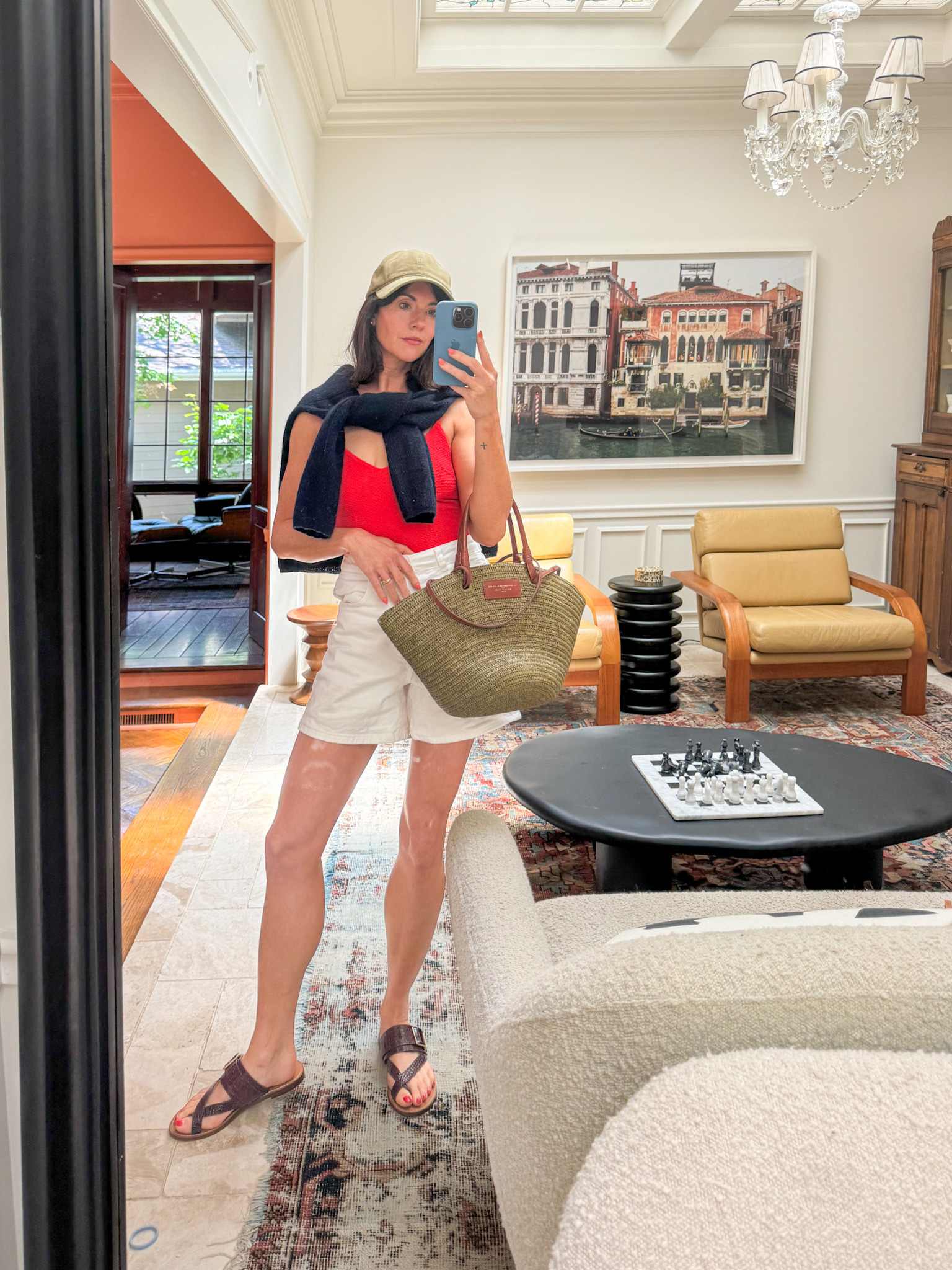 A woman wears a red swimsuit from Sézane, white high-waisted shorts, brown sandals, a straw tote bag, and a navy blue sweater draped around her shoulders