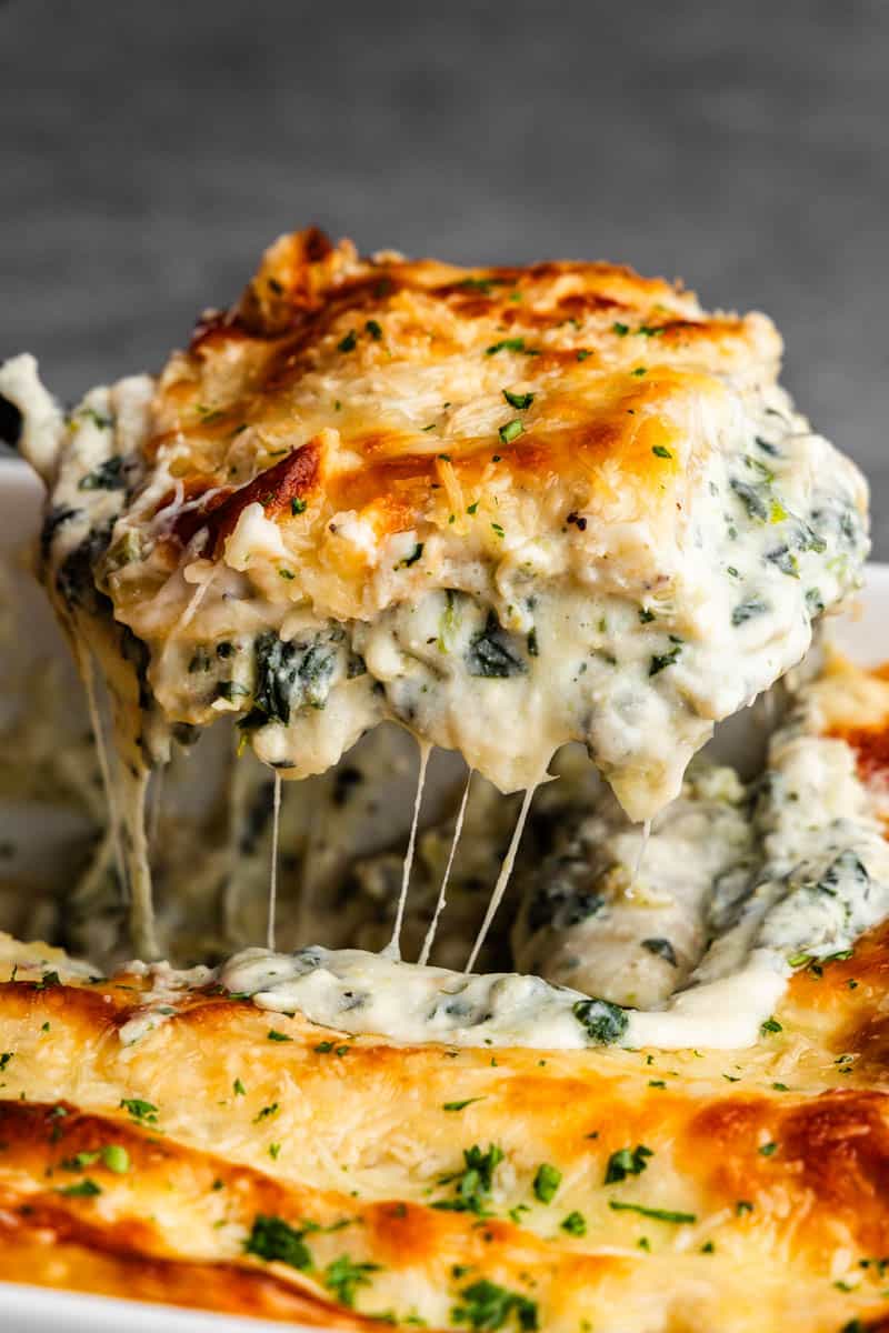 A square of white spinach artichoke lasagna being lifted from the pan with melty cheese stretching.