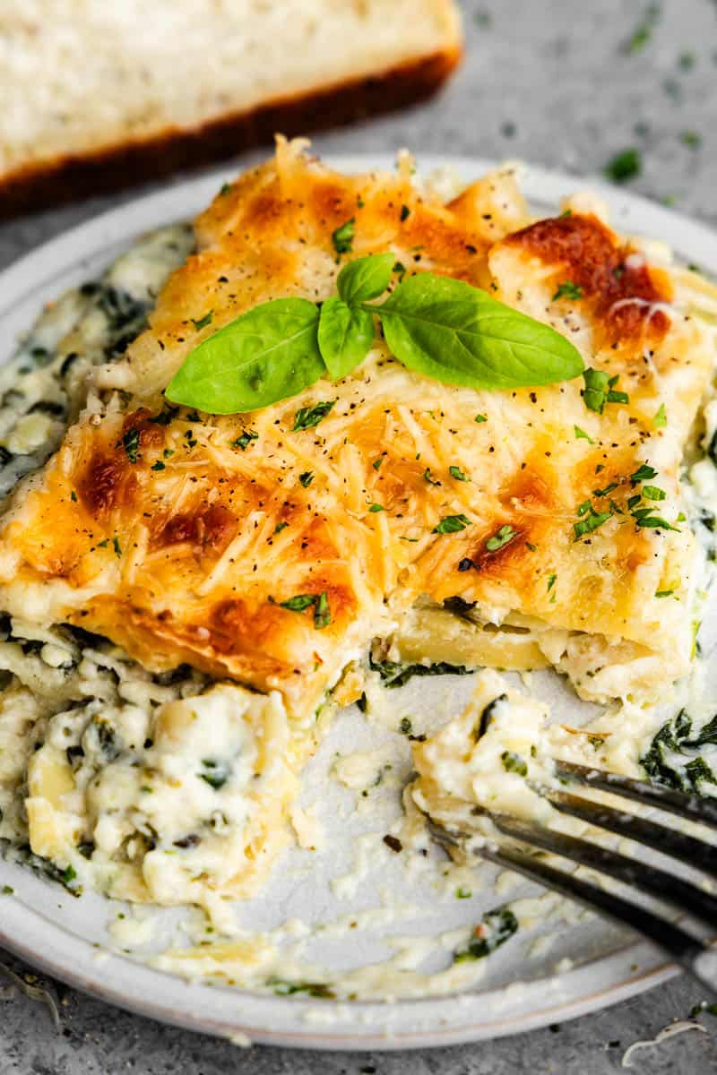 A close up view of a square of white spinach artichoke lasagna on a plate with a bite taken out of it and a fork sitting on the corner of the plate.