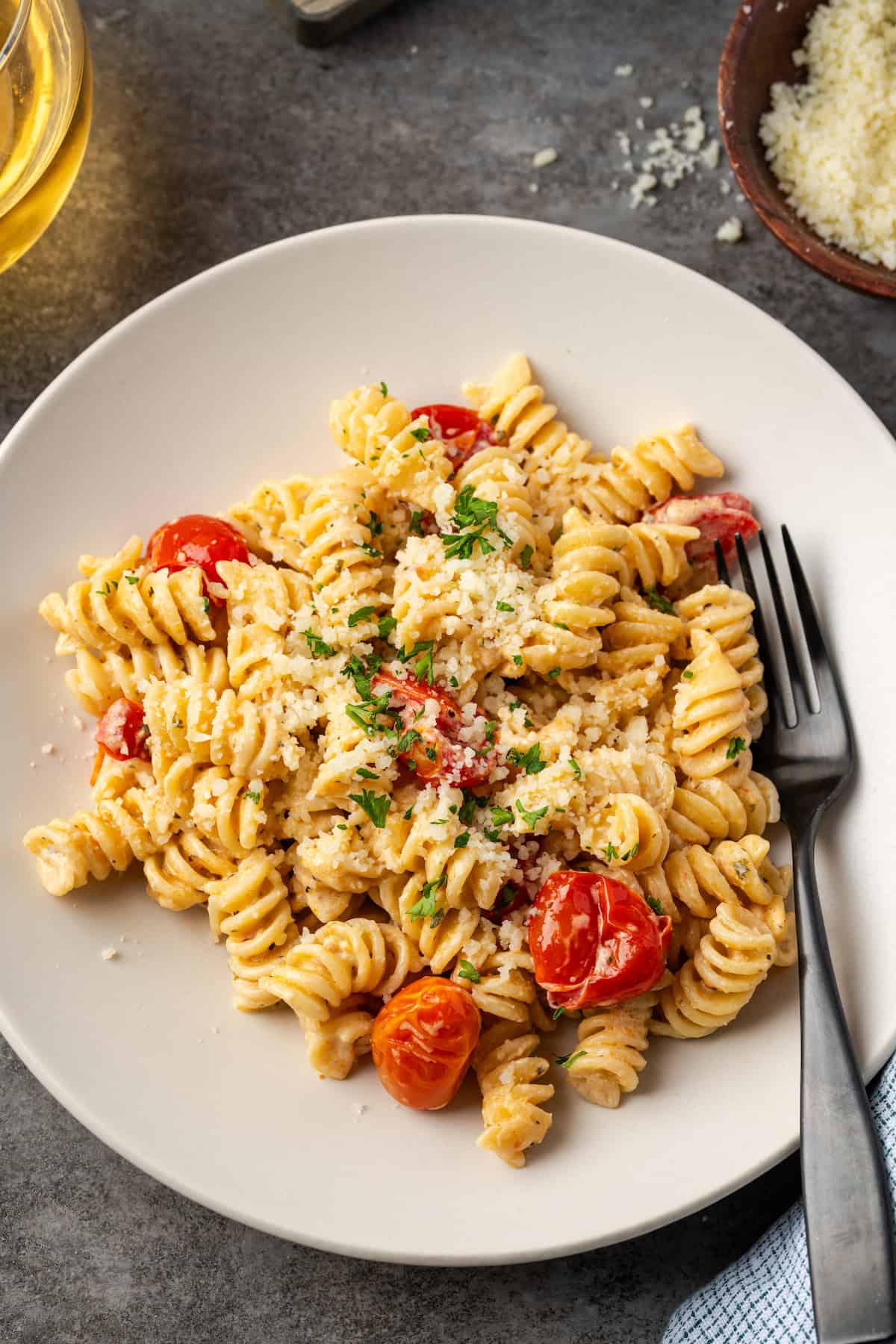 Overhead view of a fork resting in a bowl of Boursin cheese pasta, next to a small dish of parmesan and a glass of wine.