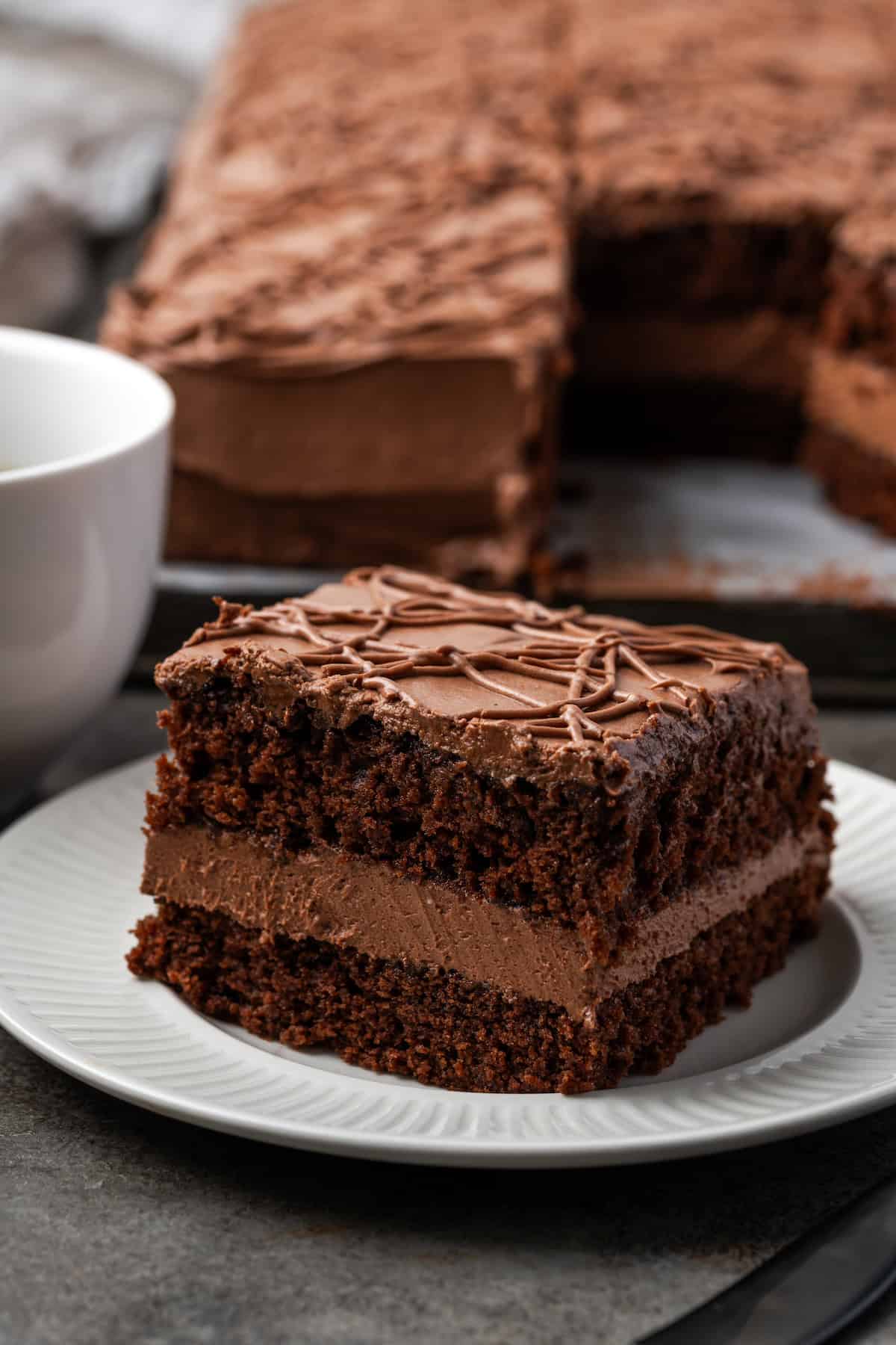 A slice of frosted fudge cake on a white plate with the rest of the cake in the background.