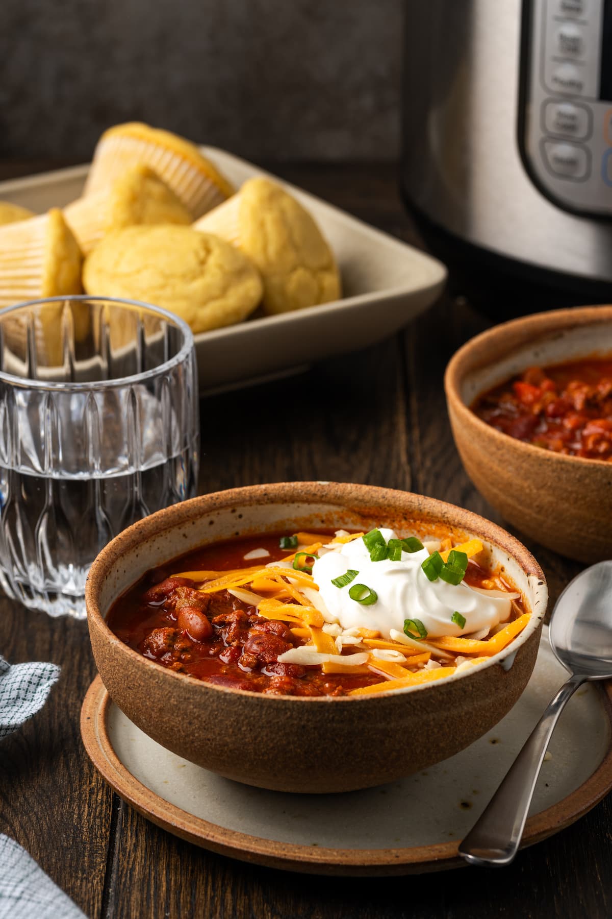 Instant pot chili garnished with shredded cheese, sour cream, and chives in a stoneware bowl, with a second bowl of chili, a plate of cornbread, and a glass of water in the background.