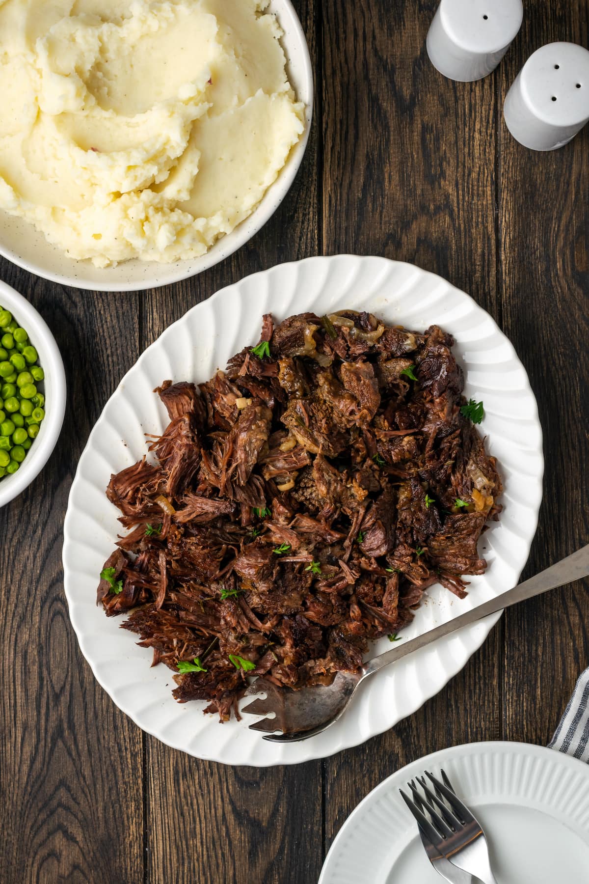 Overhead view of shredded pot roast on a white platter with a serving spoon, next to a large bowl of mashed potatoes.