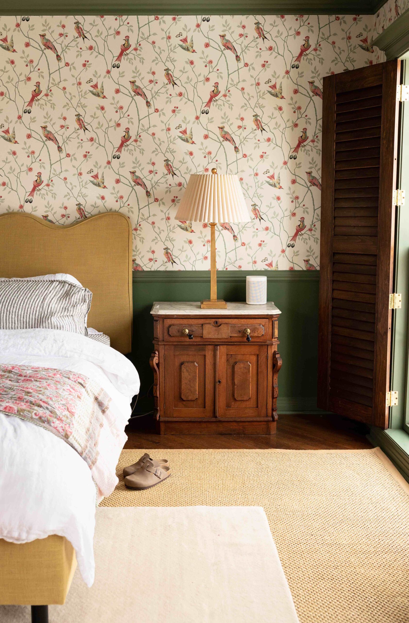 A primary bedroom with green trim, interior shutters, bird wallpaper, a scalloped headboard, antique nightstands, and layered rugs