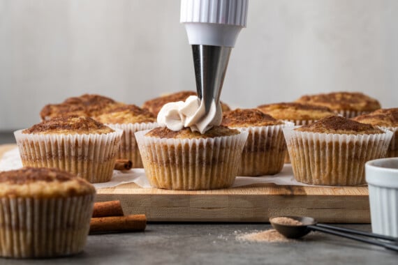 A piping tip pipes cinnamon frosting into the beginning of a swirl on a snickerdoodle cupcake, with more unfrosted cupcakes in the background.