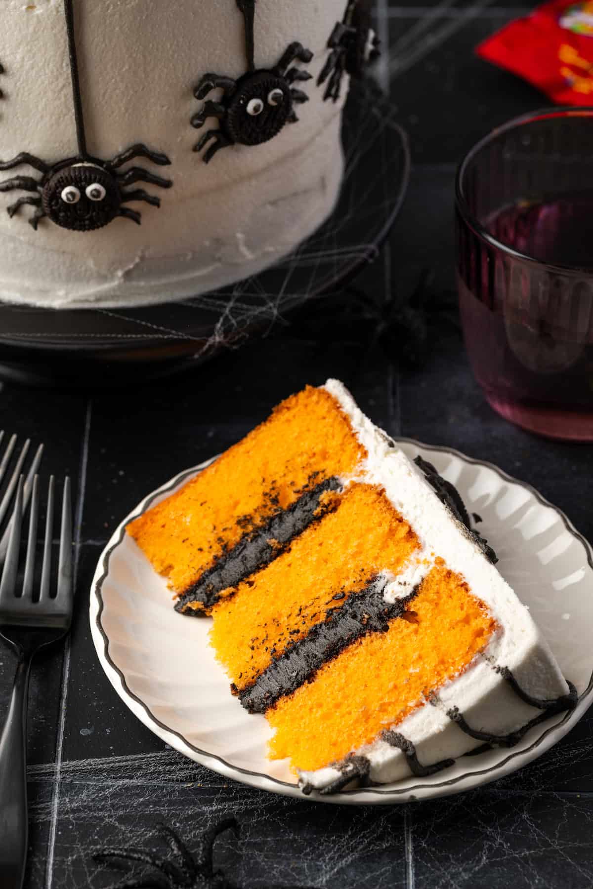 Overhead view of an orange and black spider cake on a white plate, next to the rest of the cake on a cake stand.