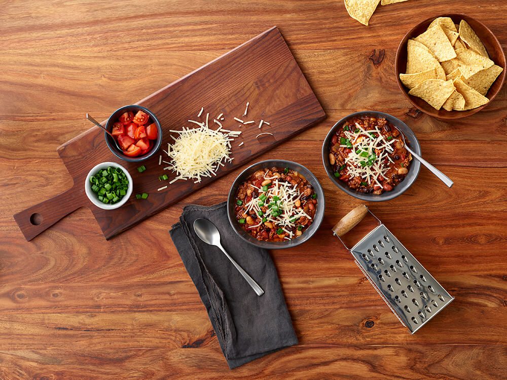 Two bowls of three bean chili sit on a wood tabletop, along with toppings like freshly grated cheese, tortilla chips, green onions, and tomatoes.