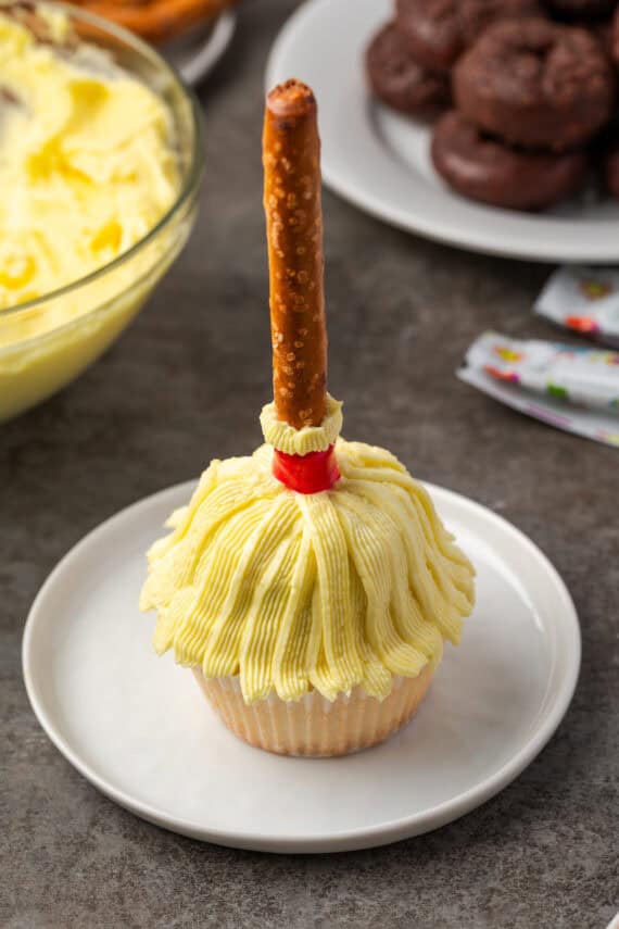 A Halloween cupcake decorated to look like a witch's broom on a white plate, with a bowl of yellow frosting and a plate of chocolate mini donuts in the background.