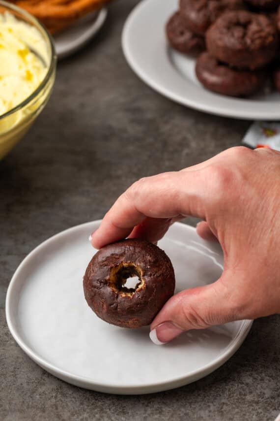 A hand holding a mini donut with the center hollowed out, over a white plate.